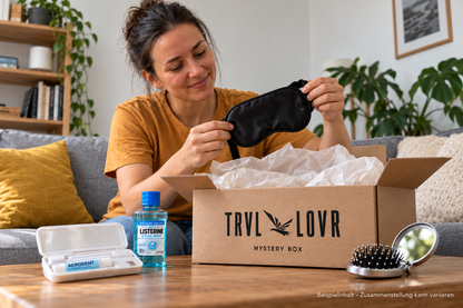 rau hält eine Schlafmaske über einer geöffneten TRVL LOVR Mystery Box mit Beispiel-Reiseutensilien